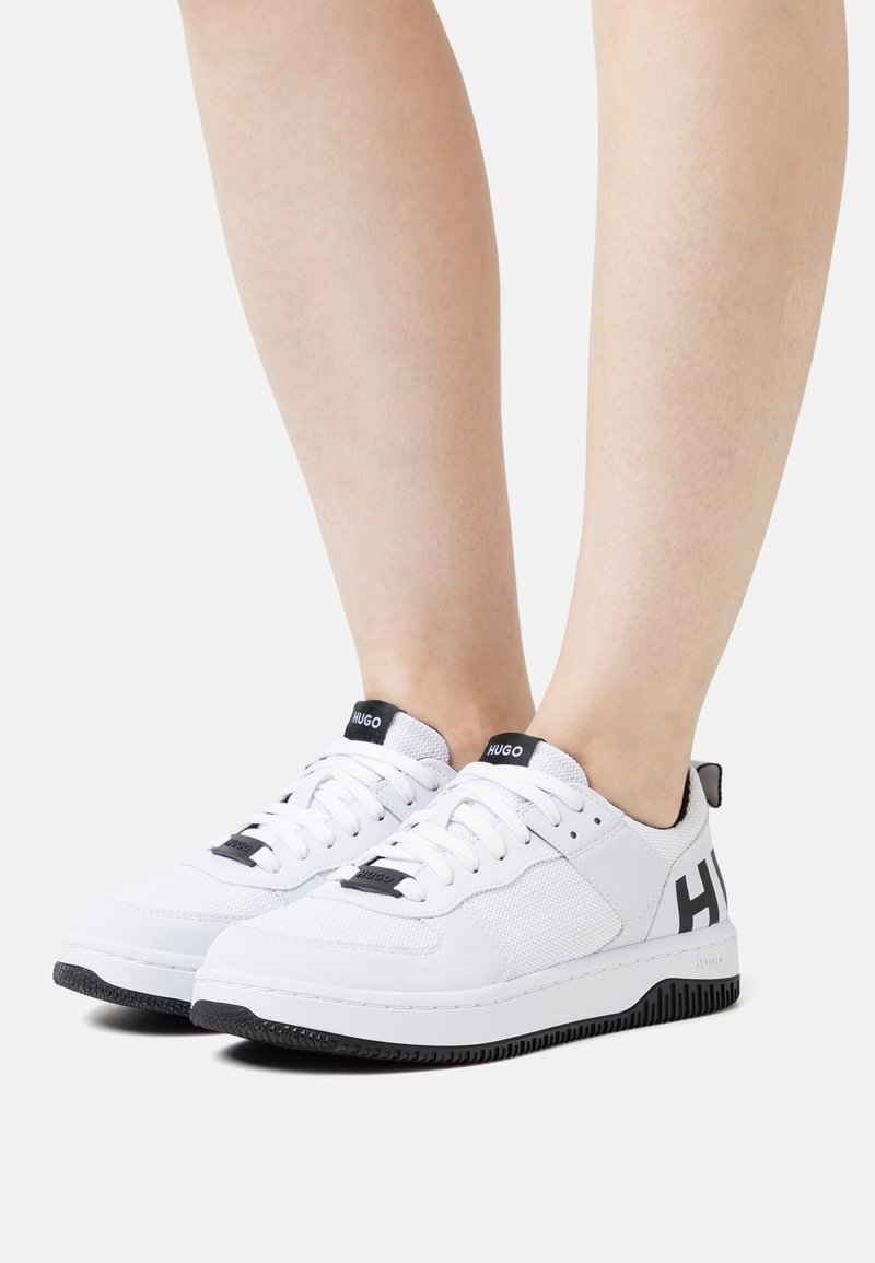 Sneakers bianchi con tomaia in rete e sintetico, dettagli neri sulla suola e sulla linguetta, con il marchio "HUGO" in evidenza. Design con punta arrotondata.