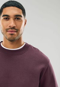 Sudadera burdeos con un escote acanalado y una textura suave. Presenta una camiseta blanca en capas, vista en primer plano de la prenda.