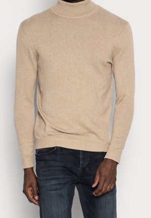Beige coltrui gedragen met donkerblauwe jeans door een persoon die staat tegen een vlakke lichte achtergrond.