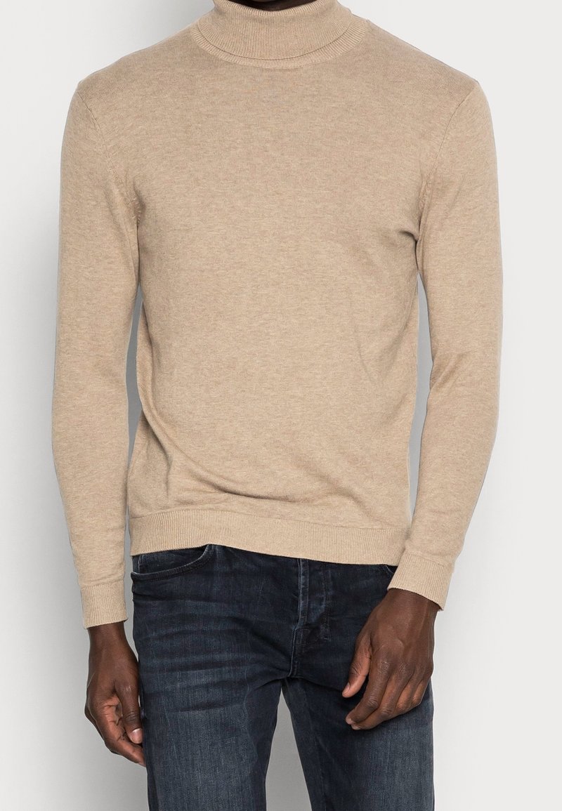 Beige polotröja bärs med mörkblå jeans på en person som står mot en enkel ljus bakgrund.