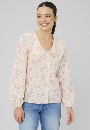 Femme souriante, portant un chemisier blanc avec imprimé floral rose, grand col, manches longues bouffantes et jean bleu sur un fond uni.
