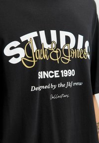 Czarna bawełniana koszulka z odważnym, kontrastującym białym i złotym napisem: "STUDIO Jack & Jones", z dodatkowymi detalami i wzorami poniżej.