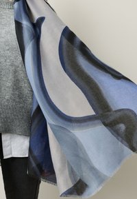 Lightweight scarf featuring abstract blue and grey waves, made from soft fabric with a frayed edge, draping elegantly from a hand.