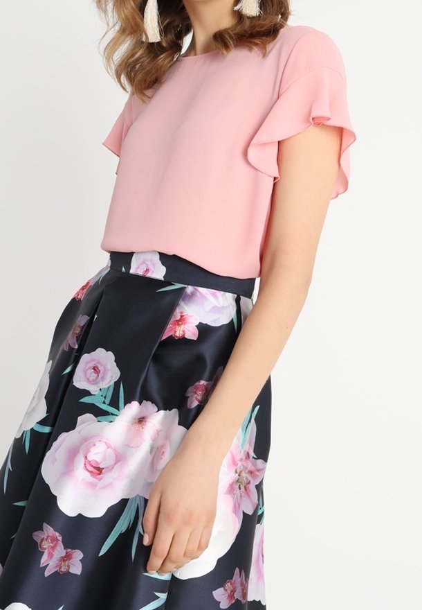 Femme portant un chemisier rose clair à manches courtes avec des manches volantées et une jupe noire avec de grands motifs floraux blancs et roses.