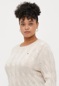 Pull en maille beige avec un col rond, doté de motifs ondulés subtils et d'un petit logo sur la poitrine. Tissu doux et texturé.