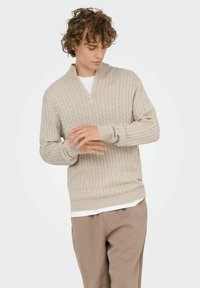 Junger Mann mit lockigem Haar, der den Ärmel eines beigefarbenen Kabelstrick-Zip-Pullovers über einem weißen Hemd anpasst, in hellbraunen Hosen.