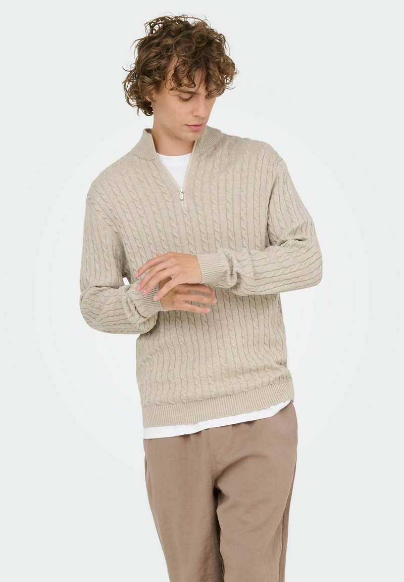 Junger Mann mit lockigem Haar, der den Ärmel eines beigefarbenen Kabelstrick-Zip-Pullovers über einem weißen Hemd anpasst, in hellbraunen Hosen.