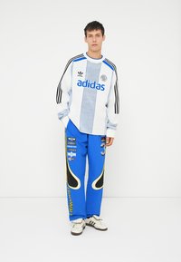 Adidasov pulover z dolgimi rokavi v beli in modri barvi z mrežastim dizajnom, v kombinaciji z modrimi trenirka hlačami z rumenimi poudarki in podrobnostmi logotipa.