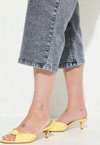Sandali con tacco in pelle gialla con un design annodato sulla parte superiore. Indossati con culotte in denim grigio, che mostrano tatuaggi alla caviglia e orlo corto.