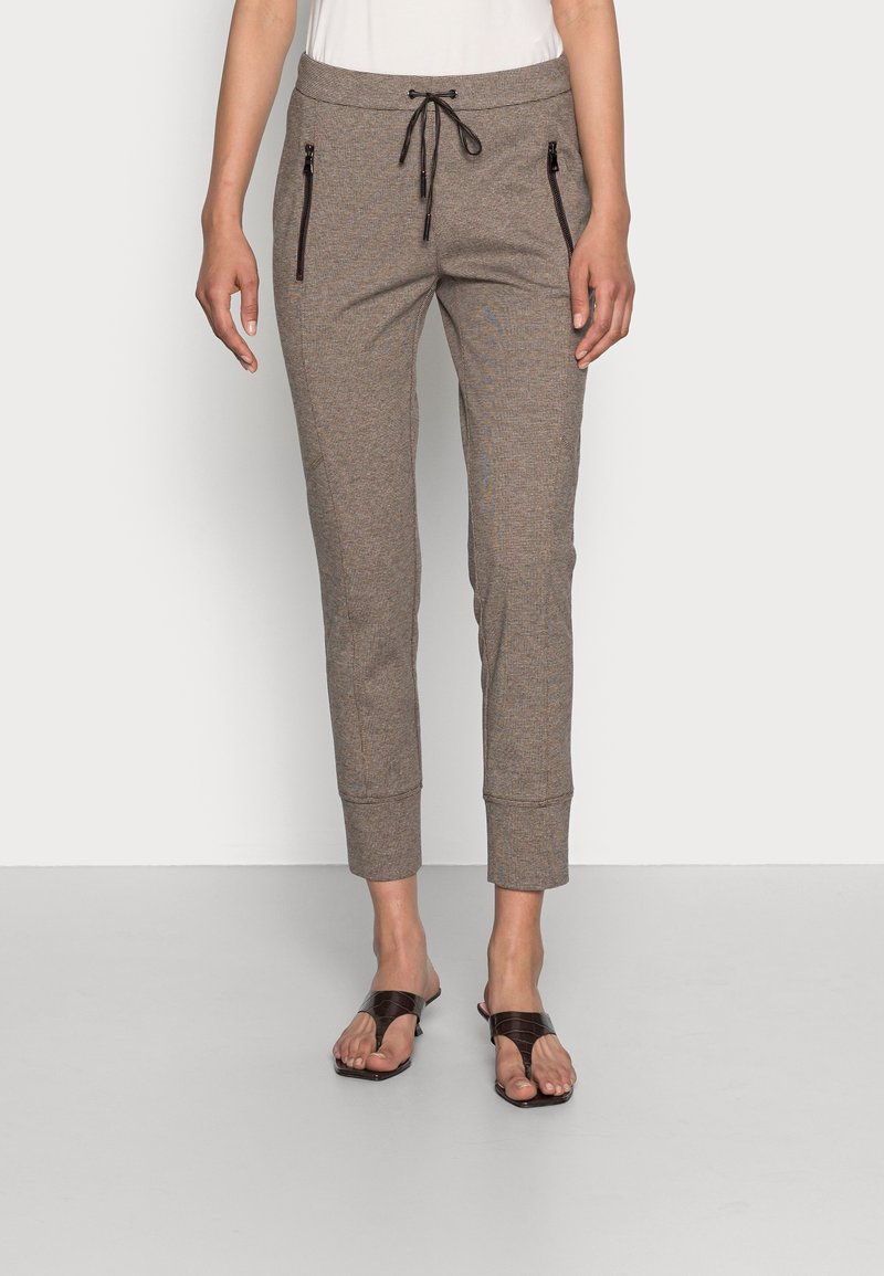 Femme portant un pantalon jogger marron fuselé avec des poches zippées à l'avant et des sandales noires à bout ouvert, debout devant un fond uni.