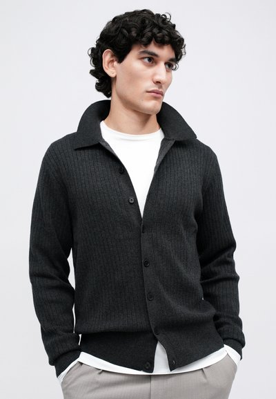 Giovane uomo con capelli ricci che indossa un cardigan a coste grigio scuro con bottoni sopra una camicia bianca e pantaloni beige chiaro, con le mani in tasca.