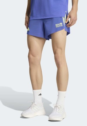 Άτομο που φορά μπλε σορτς BMW Berlin Marathon, λευκές κάλτσες Adidas και λευκά αθλητικά παπούτσια, στέκεται σε ανοιχτό φόντο.