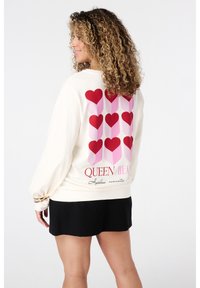Vrouw in een crèmekleurige sweatshirt met rode en roze hartkolommen en de tekst "Koningin van Harten", staande in profiel tegen een witte achtergrond en met een zwarte rok.