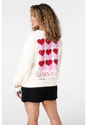 Femme portant un sweat-shirt crème avec des colonnes de cœurs rouges et roses et le texte "Reine des Cœurs", et une jupe noire, se tenant de côté contre un fond blanc.