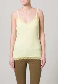 Gul ribbad linne med spetsdetaljer vid halsringning och nederkant; justerbara spaghetti axelband. Matcher med bruna slim-fit byxor.