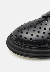 Chaussure en cuir noir perforé à bout arrondi, finition lisse et semelle en caoutchouc épaisse. Présente un accent discret de couture le long du bord.