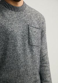 Hombre vistiendo un suéter de punto gris con un cuello acanalado y un bolsillo cuadrado en el pecho, sobre un fondo claro y liso.