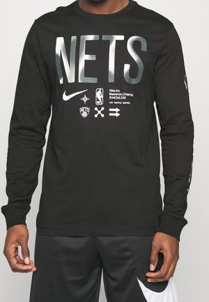 Chemise noire à manches longues avec texte argenté "NETS", logos Nike et NBA, ainsi que d'autres petits graphismes à l'avant, portée avec un short noir et blanc.