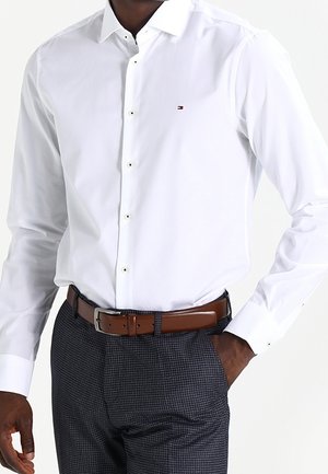 Mężczyzna w dopasowanej białej koszuli zapinanej na guziki wpuszczonej w ciemne spodnie w kratę z brązowym skórzanym paskiem, ręka w kieszeni.