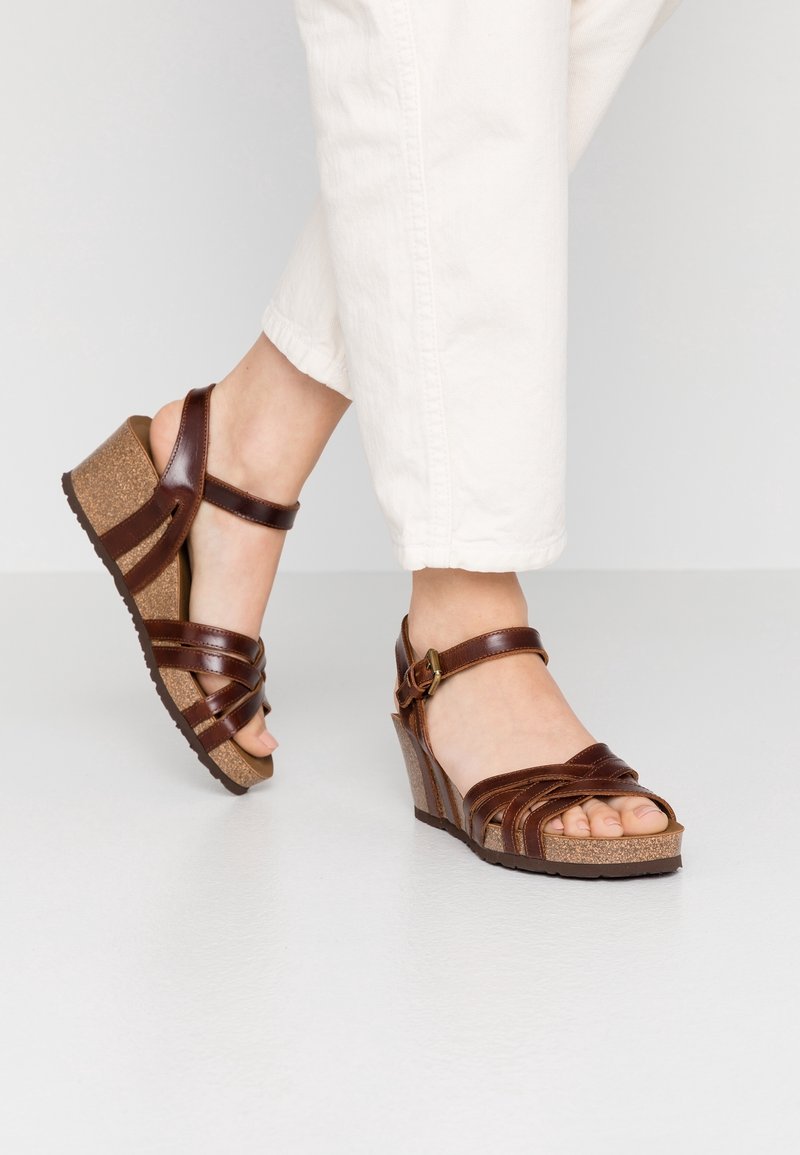 Pieds en pantalon blanc portant des sandales compensées en cuir marron avec semelles en liège sur un fond clair uni.