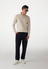 Cămașă polo bej cu mânecă lungă și două butoane, asociată cu pantaloni bleumarin și adidași albi. Ochelarii de soare completează ținuta.