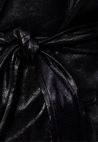 Czarna skórzana faktura z wzorem wężowym, z centralnym węzłem i opadającymi drapowaniami, podkreślająca błyszczące wykończenie.