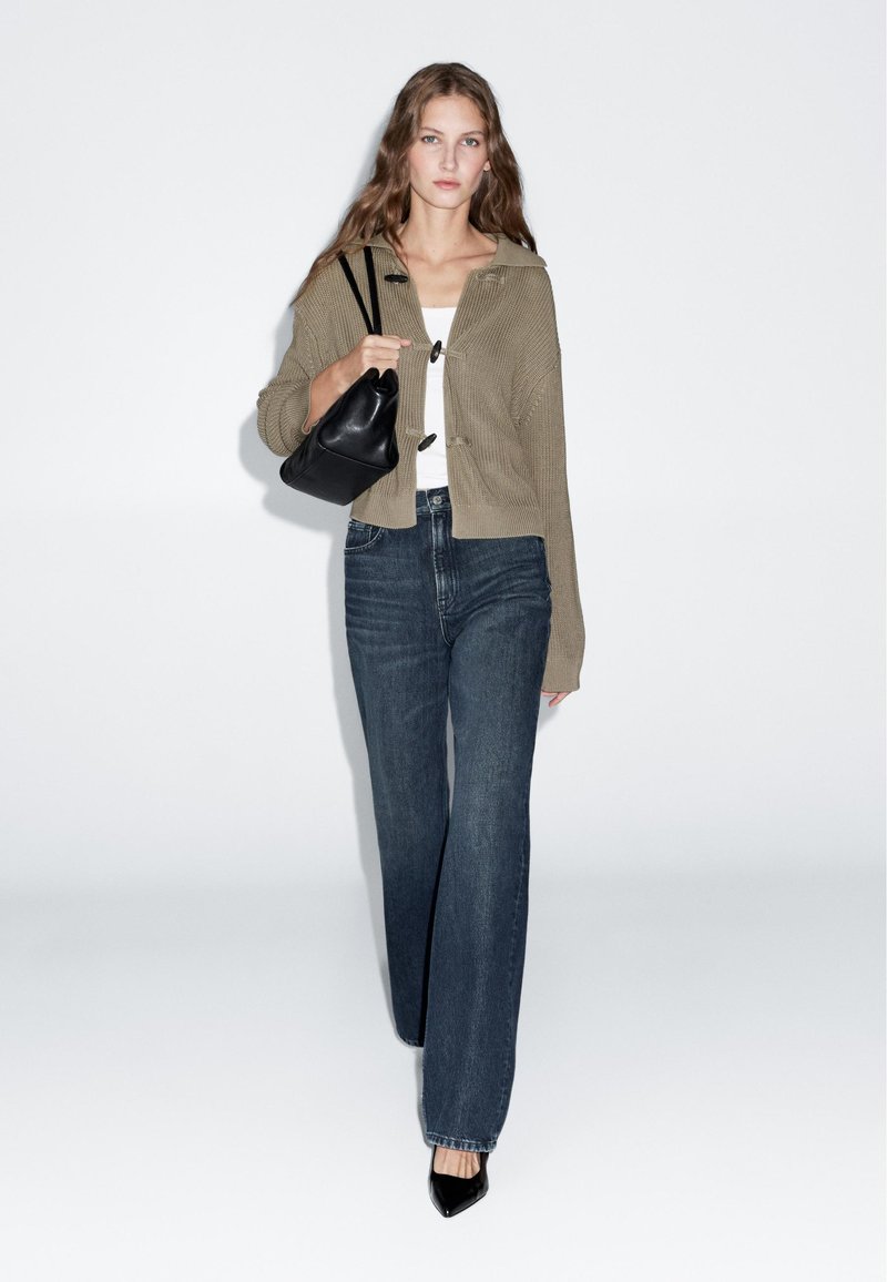 Gebreid grijs-bruin cropped vest met capuchon, zwarte handtas, wit topje en blauwe wijde broekspijpen jeans. Zwarte hakken maken de outfit compleet.