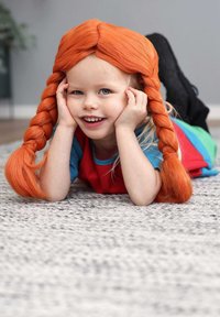 Barn med lang orange fletning, iført et farverigt sæt med blå og røde detaljer, liggende på et tekstureret gråt tæppe.