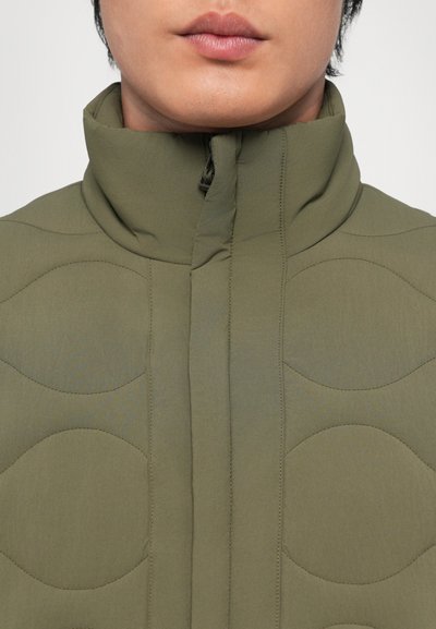 Jachetă din materiale umplute de culoare verde măslin, cu guler înalt, care prezintă modele circulare matlasate și un zipper central pentru închidere. Material moale și texturat.