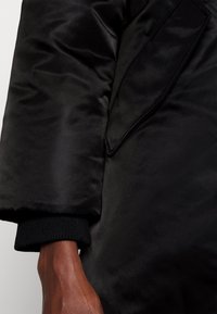 Giacca imbottita nera con texture lucida, polsini neri a costine e dettaglio di tasca piatta sul braccio. Vista ravvicinata della manica e della mano.