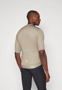 Kortermet, lys tan atletisk topp med et tettsittende design, teksturert stoff og en baklomme. Har logo og tekst nær sømmen.