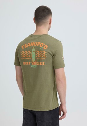 Mand iført olivengrøn T-shirt med orange og grøn surfing-tema grafik og teksten "TEAHUPO'O REEF BREAKS" på ryggen.