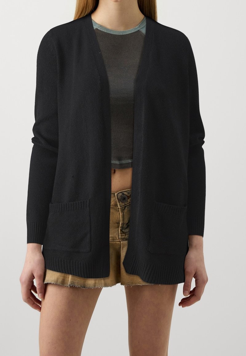 Femme portant un cardigan noir ouvert avec des poches sur un haut court foncé et un short taille haute marron clair.