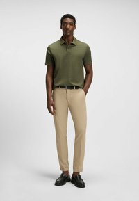 Koszula polo krótki rękaw w kolorze oliwkowym z teksturowanym wzorem, w połączeniu z jasnobeżowymi spodniami i czarnymi loafersami. Mężczyzna stoi z rękami w kieszeniach.