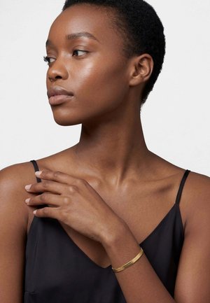 Lyhythiuksinen nainen, joka pukeutuu mustaan hihattomaan topiin ja kultaiseen rannekoruun, katsoo vasemmalleen käden lepääessä olkapäällä.