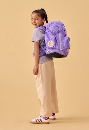 Junges Mädchen lächelt, trägt ein lila Shirt und beige Hosen und hat einen großen lila Rucksack mit weißen Punkten und Cartoon-Aufnähern dabei.