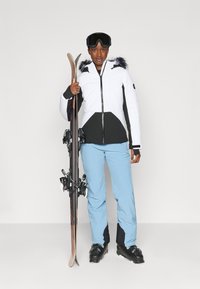 Weiße und schwarze isolierte Skijacke mit Pelzbesatz, kombiniert mit hellblauen Skihosen und schwarzen Skischuhen. Skis in der linken Hand gehalten.
