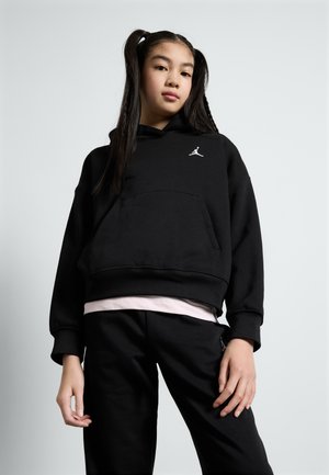 Sort hættetrøje i blød bomuld med en kængurulomme og hvidt logo på brystet, parret med sorte sweatpants, der har en lagdelt lyserød skjorte under.