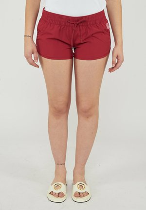 Donna con pantaloncini rossi con coulisse, sandali bianchi a ciabatta e top bianco, in piedi davanti a uno sfondo chiaro e semplice.