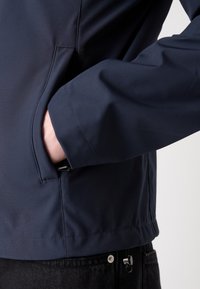 Temno modra jakna z gladko površino, ki ima stransko žep in kontrastno šivanje. Poudarjen detajl rokava in žepa.