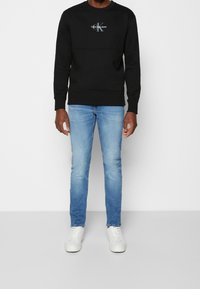 Svart sweatshirt med broderad logotyp, kombinerad med ljusblå jeans. Skorna är vita sneakers. Båda plaggen har en avslappnad passform.
