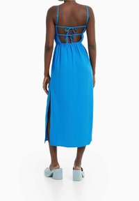 Žena v modrých bezramínkových midi šatech s otevřeným zádovým výstřihem a šňůrkami, doplněná světlomodrými sandály na podpatku, stojící na bílém pozadí.