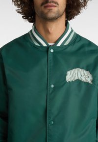 Vihreä, sileä kangastakki, jossa on napilla suljettava etuosuus, raidallinen kaula-aukko ja vaaleansinisellä brodeerattu "VANS"-logo.