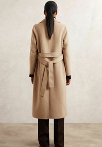 Palton bej din lână, cu talie cu brâu, mâneci lungi și guler cu decupaje. Textură netedă și design de lungime medie. Pantaloni maro vizibili dedesubt.