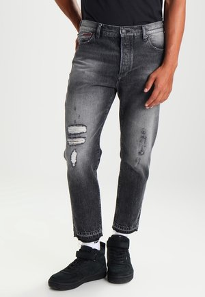 Mann trägt schwarze, abgenutzte Jeans, weiße Socken und schwarze High-Top-Sneaker, steht vor einem einfarbigen hellen Hintergrund.