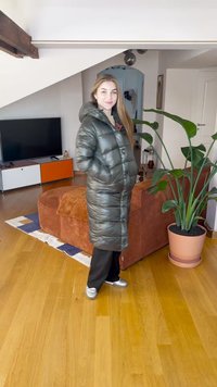 Ilgas blizgus žalias pūkinis paltas su gaubtu, priekyje užtrauktuku ir šoninėmis kišenėmis. Dėvimas ant juodų kelnių ir sidabrinių sportinių batelių, viduje, šalia augalų.