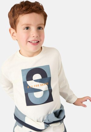 T-shirt a maniche lunghe color crema con una grafica blu e nera che presenta la scritta "S IS FOR SKATE", indossato con accenti blu legati attorno alla vita.