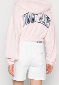 Roze sweatvest met capuchon en "TOMMY JEANS" geborduurd op de achterkant, gecombineerd met witte denim shorts voorzien van achterzakken en een logolabel.