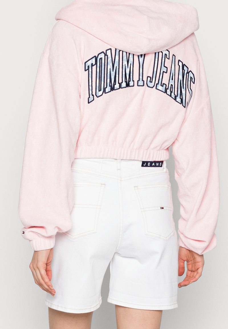 Roze sweatvest met capuchon en "TOMMY JEANS" geborduurd op de achterkant, gecombineerd met witte denim shorts voorzien van achterzakken en een logolabel.