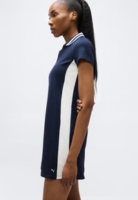 Γυναίκα που στέκεται πλαγίως φορώντας ένα μπλε σκούρο φόρεμα πόλο με λευκές πλευρικές πινελιές και ριγωτό γιακά, με μακριά σκούρα μαλλιά σε αλογοουρά.